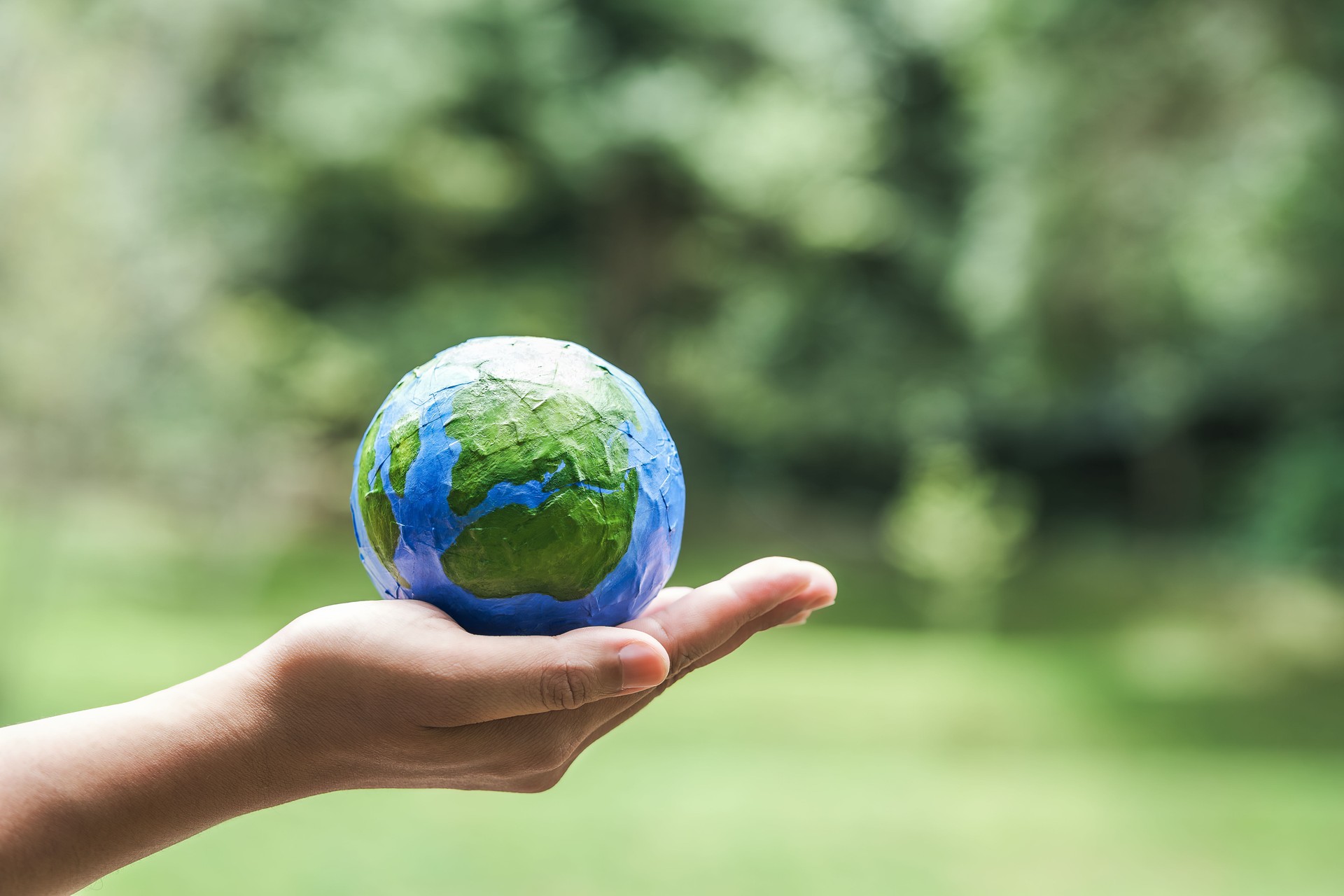 Hand holding a painted paper model of Earth with green natural background, symbolizing environmental care, sustainability, ecology, and global responsibility.